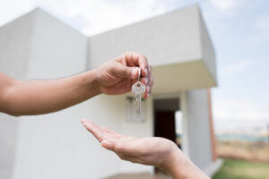 Uma mão entregando chave numa outra mão com uma casa branca ao fundo