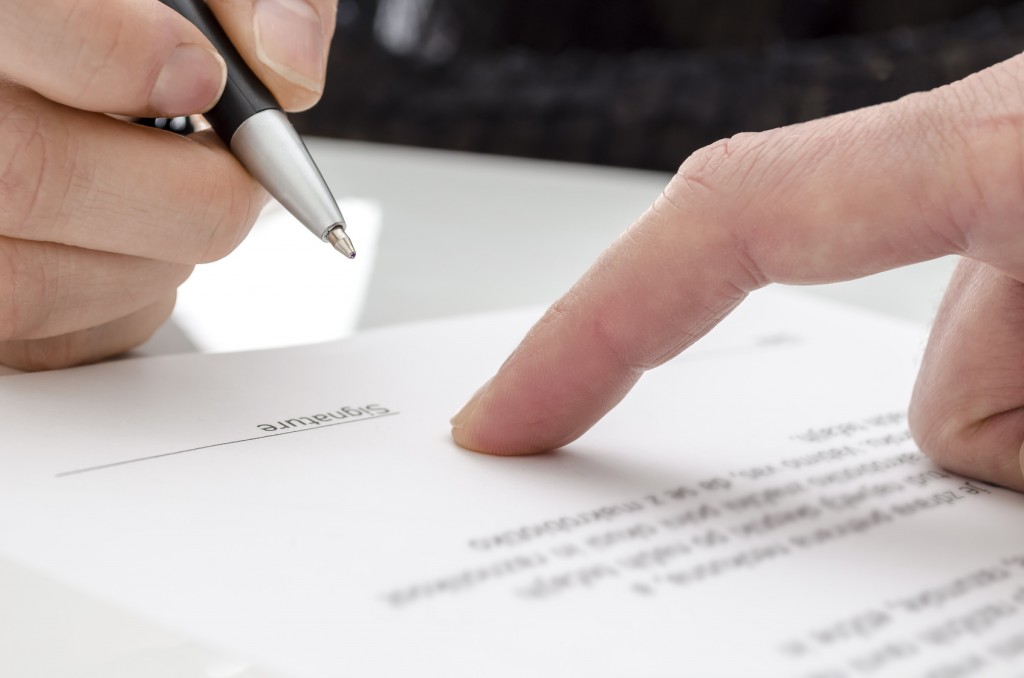 Uma mão apontando uma cláusula de um contrato em cima de uma mesa e, do outro lado, uma mão segurando uma caneta preta para assinar este contrato