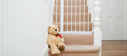 Urso de pelúcia bege, grande, com cachecol vermelho no pescoço, sentado no último degrau de uma escada e acima dele um portão pequeno impedindo a passagem pela escada