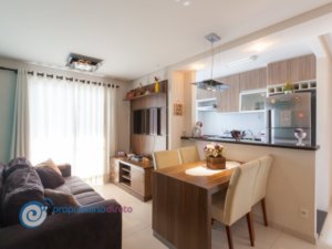 Sala de apartamento com cozinha americana, mesa retangular em madeira de quatro lugares e sofá marrom.