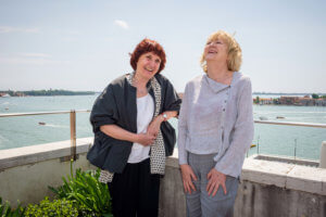 Duas mulheres sorrindo, próximas ao parapeito de uma passarela, onde atrás se vê um rio com alguns barcos