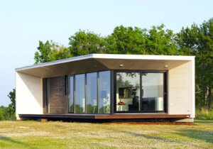 Pequena casa modular, com paredes brancas e grandes janelas de vidro, sobre um gramado